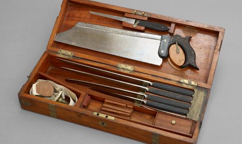 An amputation set, featuring various sharpened tools of different lengths and sizes including a large metal saw, stored in a wooden box with allocated compartments. 