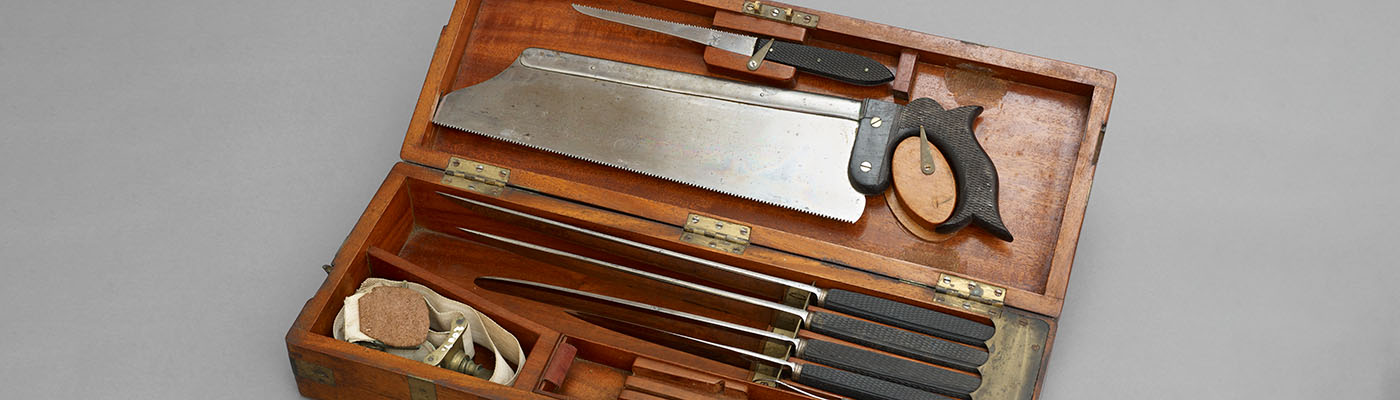 An amputation set, featuring various sharpened tools of different lengths and sizes including a large metal saw, stored in a wooden box with allocated compartments.