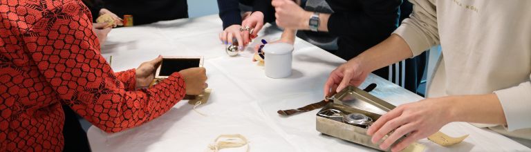  A group of MA Art Gallery and Museum Studies examine, handle and photograph museum objects and instruments laid out on a cloth-covered table.