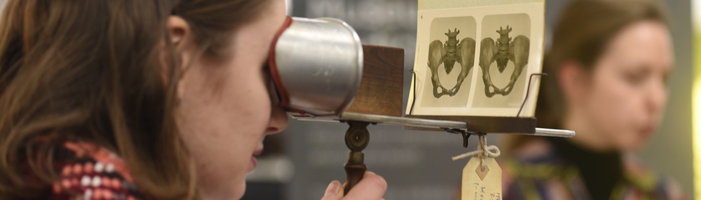 A young woman looks through a stereoscopic viewer, an anatomical teaching device. She examines a 3D image of the bone structure of an enlarged female pelvis, printed on a viewing card. 