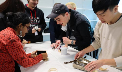  A group of MA Art Gallery and Museum Studies examine, handle and photograph museum objects and instruments laid out on a cloth-covered table.