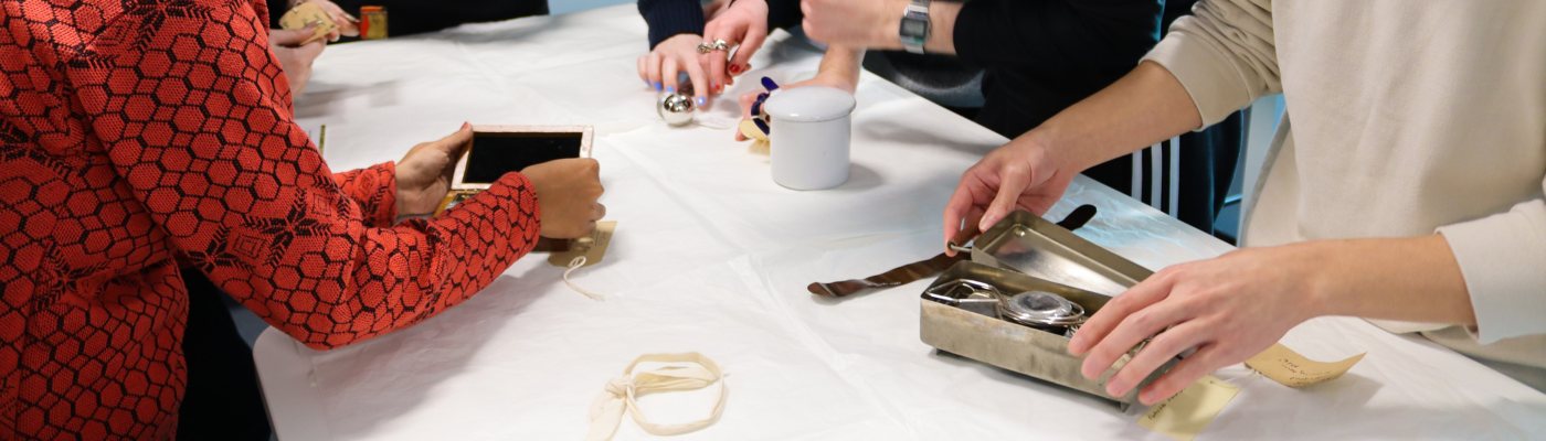 A group of MA Art Gallery and Museum Studies examine, handle and photograph museum objects and instruments laid out on a cloth-covered table. 